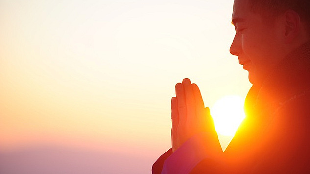 a person with eyes closed and hands in a prayer posture with sun in the background