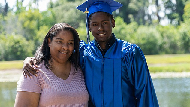 a young black man, wearing his graduation cap and robe, has his arm around his smiling mother's shoulders