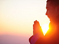 a person with eyes closed and hands in a prayer posture with sun in the background