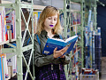 a young woman standing by a bookshelf reading a book