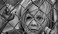 a sad child standing behind a chain link fence and holding on to the wire as he looks out beyond the fence