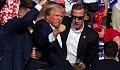 Trump with blood on his face holds up a clenched fist while people in suits surround him, illustrating the impact of social media on political violence.